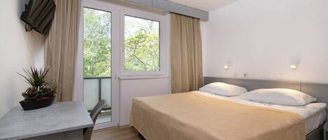 A bedroom with a bed, a desk, a potted plant, a window with curtains, and a view of trees.