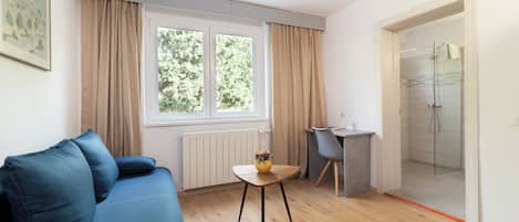 A modern living room with a blue sofa, a small wooden table with a vase, a glass shower enclosure, and a view of greenery outside.