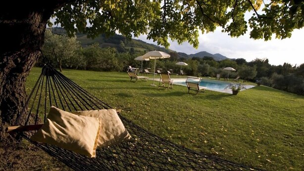 Una hamaca bajo un árbol, una piscina y mobiliario exterior con sombrillas.