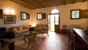 Una sala de estar acogedora con un sofá, sillas, una mesa de centro y una mesa de comedor. Hay ventanas y una puerta que da al exterior.