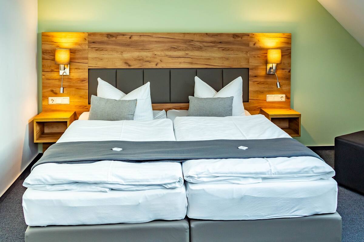 Een hotelkamer met een houten hoofdeinde, twee nachtkastjes met lampen, een bed met wit beddengoed en een grijze bank.