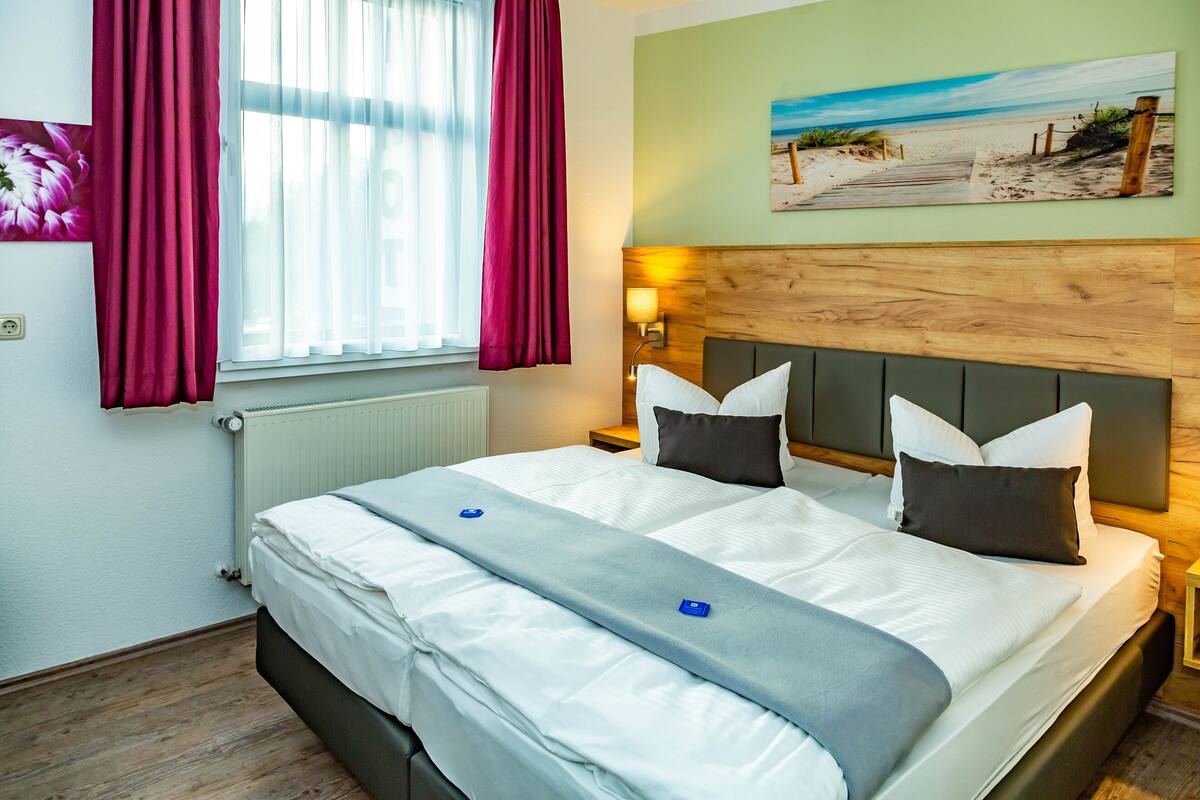 Een netjes opgemaakt bed met witte lakens en een grijze plaid, een houten hoofdbord, een nachtkastje met een lamp, en een schilderij met een strandthema aan de muur.