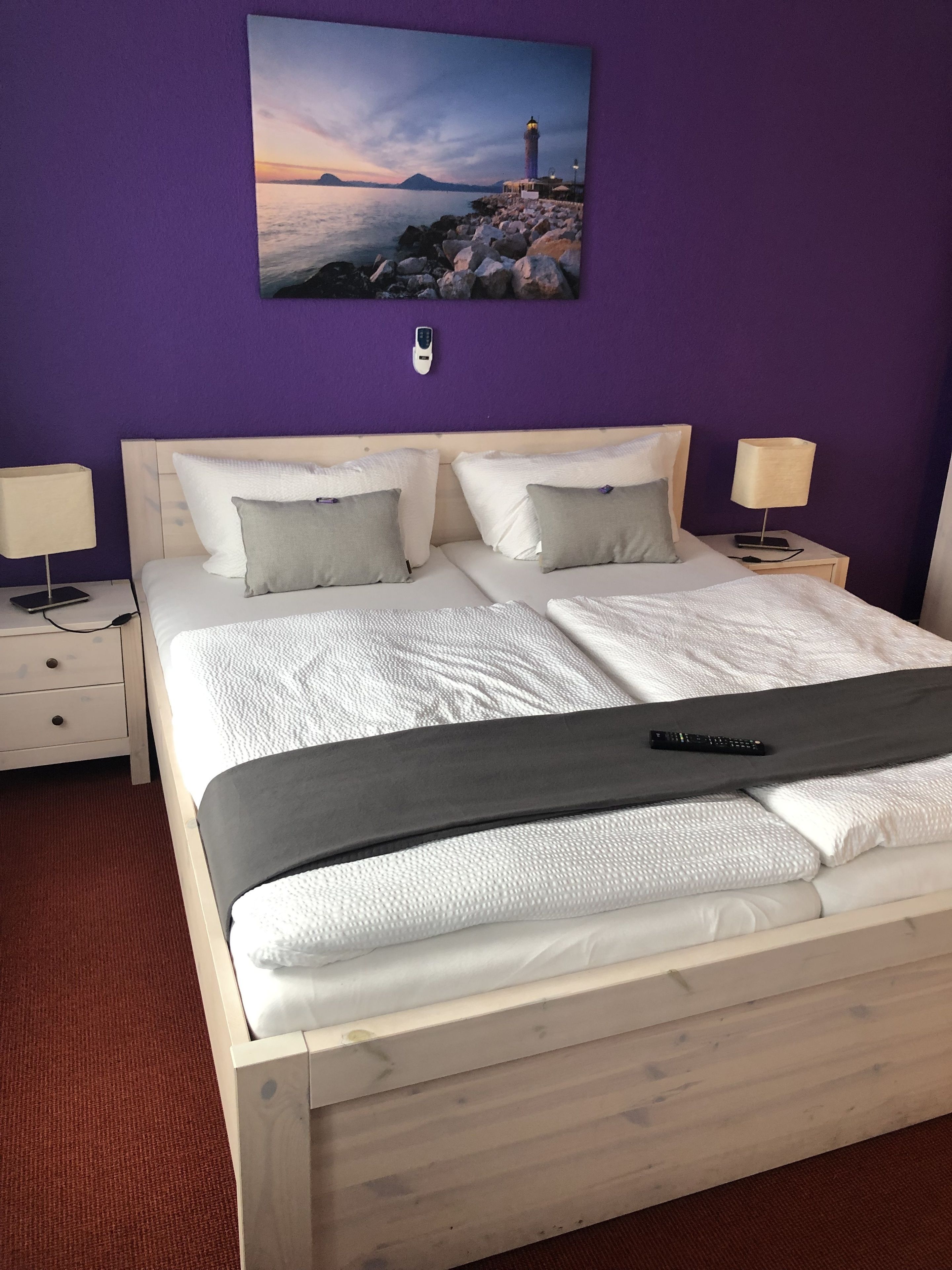 A bedroom with a bed, two bedside tables, a nightstand, a wall-mounted phone, and a painting of a lighthouse.