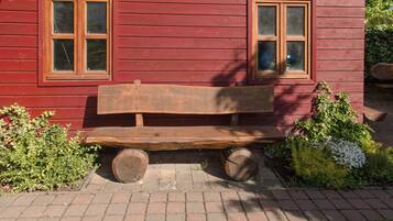 Un banc en bois, dont les roues sont constituées de deux grosses bûches, est placé devant un bâtiment en bois rouge doté de deux fenêtres.