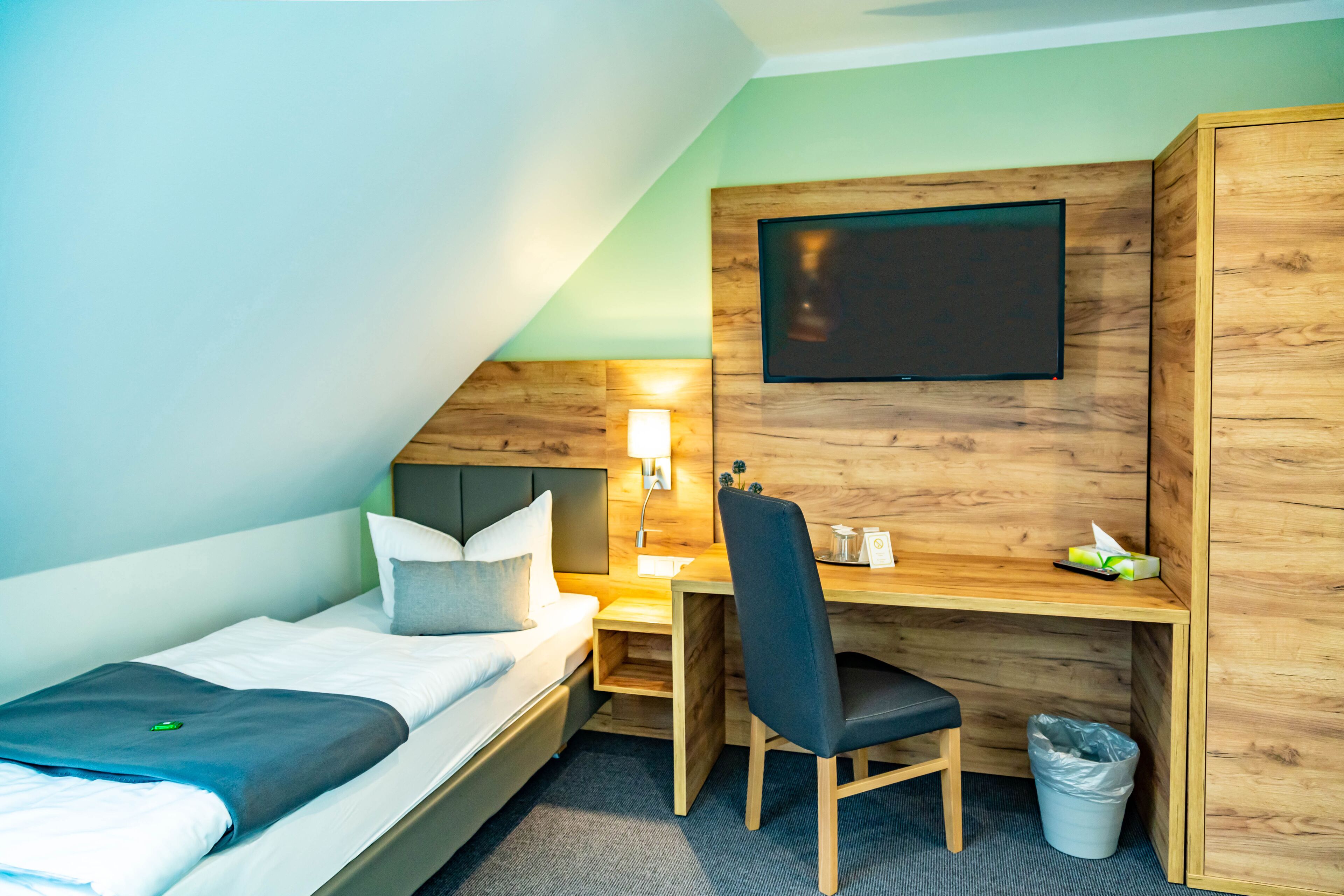 Habitación de hotel pequeña con una cama individual, un escritorio, una silla, un televisor montado en la pared y un cesto de basura.