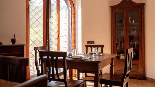 A dining area with a wooden table and chairs, a cabinet with glass doors, and a large arched window. - Villetta Capodimonte Guest House (Naples)