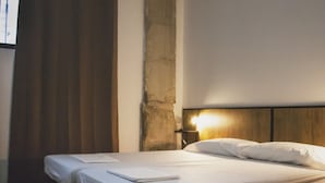 Une chambre d’hôtel avec deux lits simples, une tête de lit en bois, une table de chevet avec une lampe et une grande fenêtre avec des rideaux.