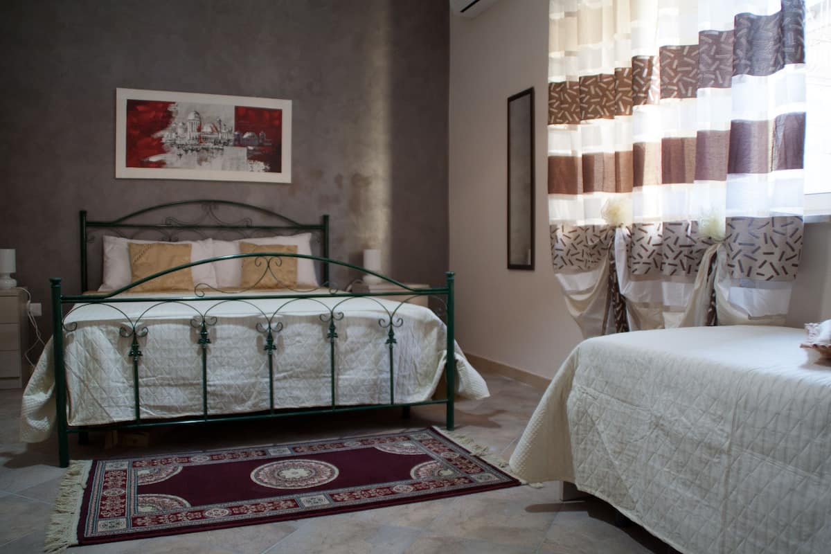 Une chambre avec un lit en métal vert, une couvre-lit blanche matelassée, un tapis rouge à motifs et un tableau représentant un bâtiment accroché au mur.