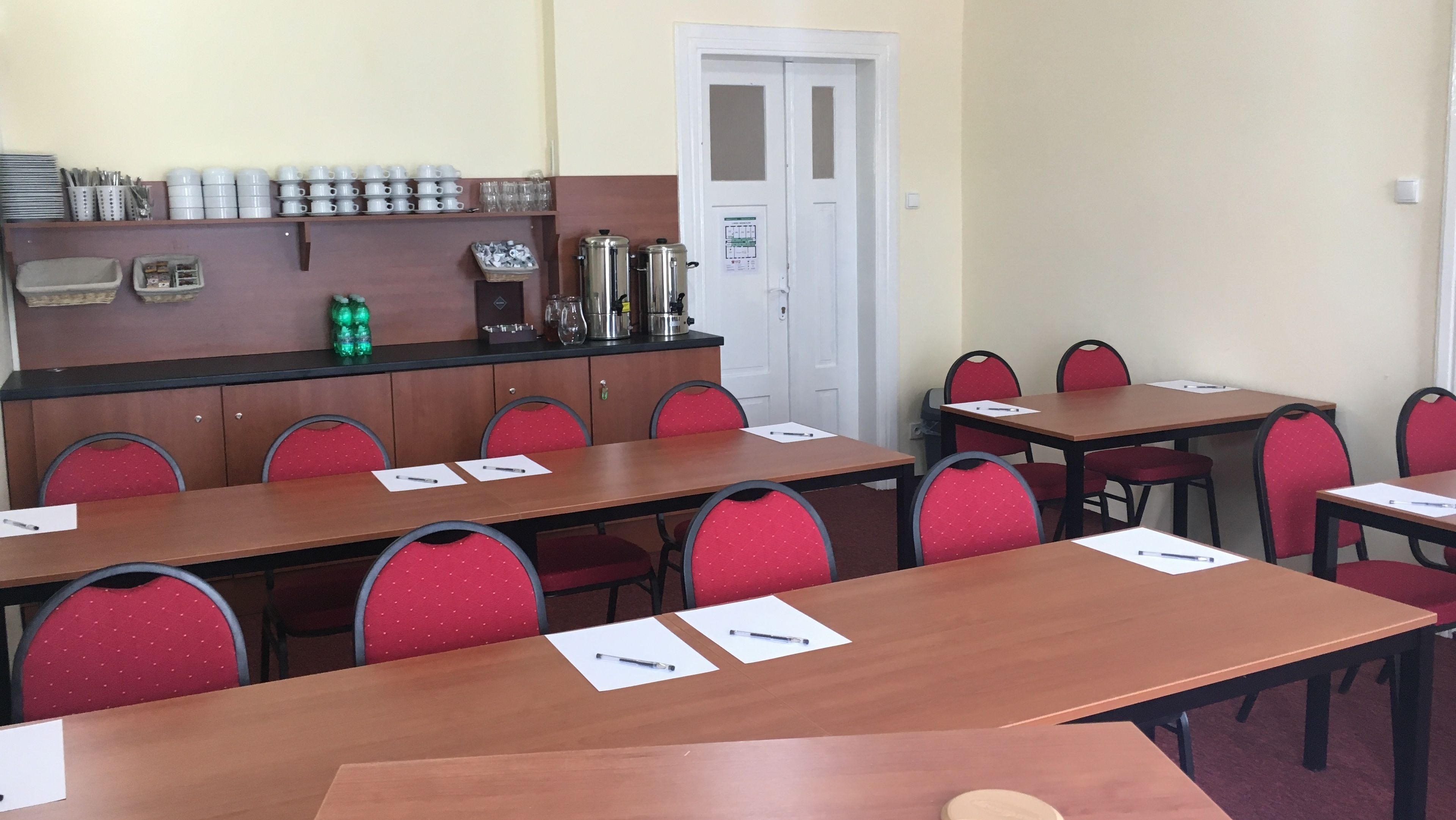 Una sala conferenze con tavoli in legno e sedie rosse, dotata di penne e carta, e un angolo cottura con macchine da caffè e contenitori portaoggetti.