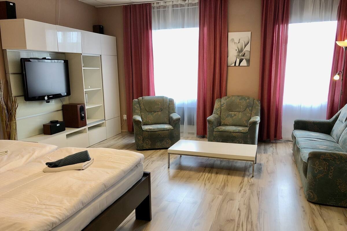 Ett hotellrum med en säng, två fåtöljer, en soffa, en platt-TV, en bokhylla och en tavla på väggen.
