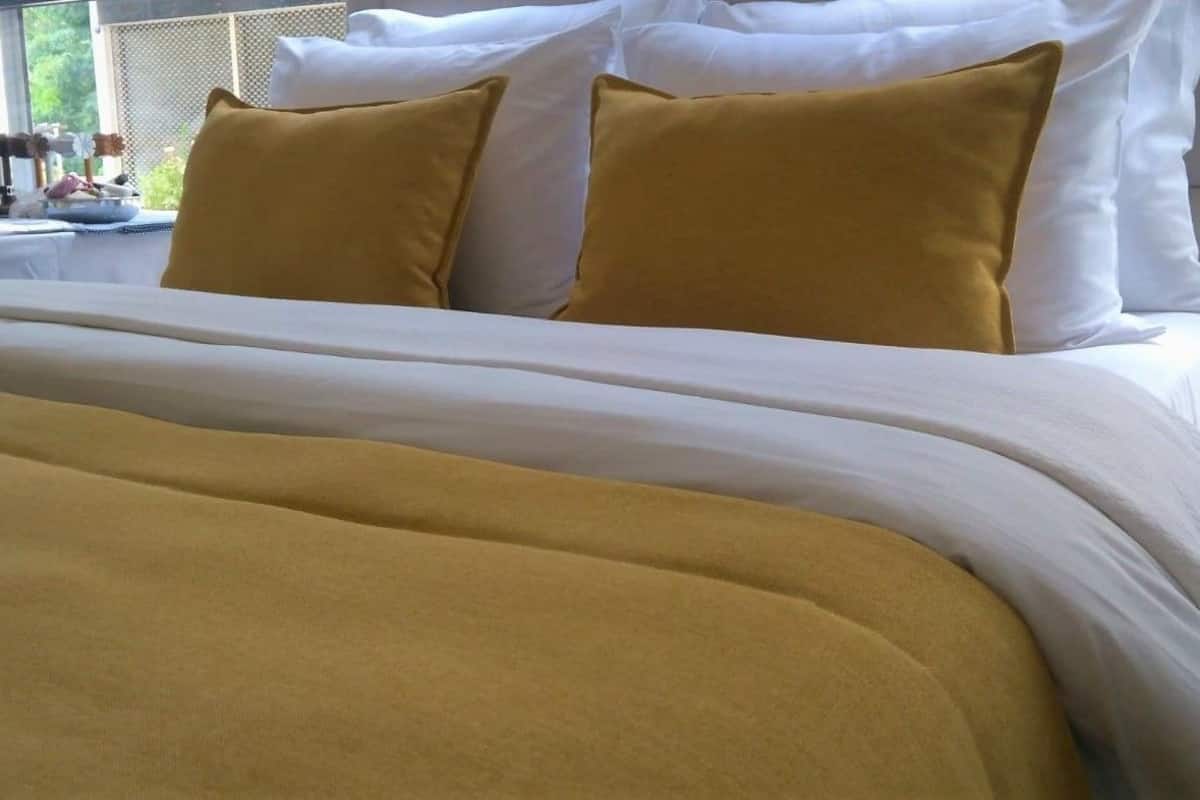 A bed with white and mustard-colored bedding, a window with a view of greenery, and a table with items on it.