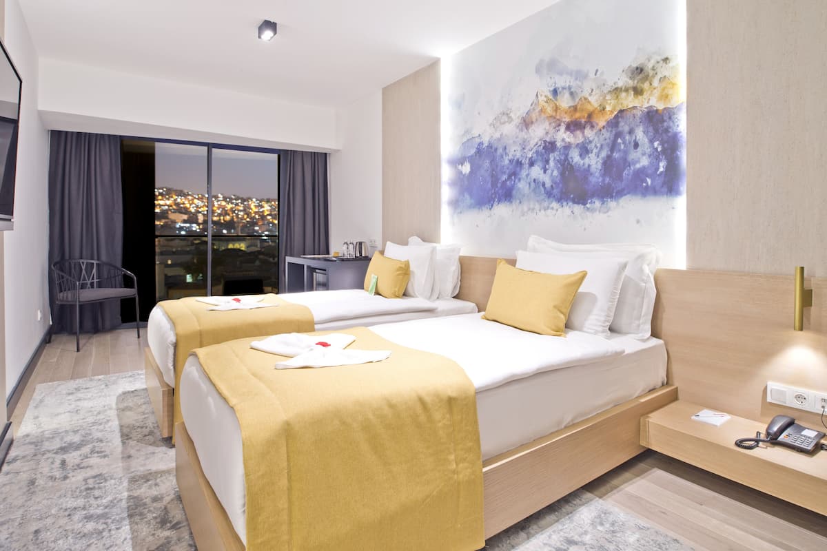 A hotel room with two beds, a desk, a chair, a large abstract painting, and a city view through the window.