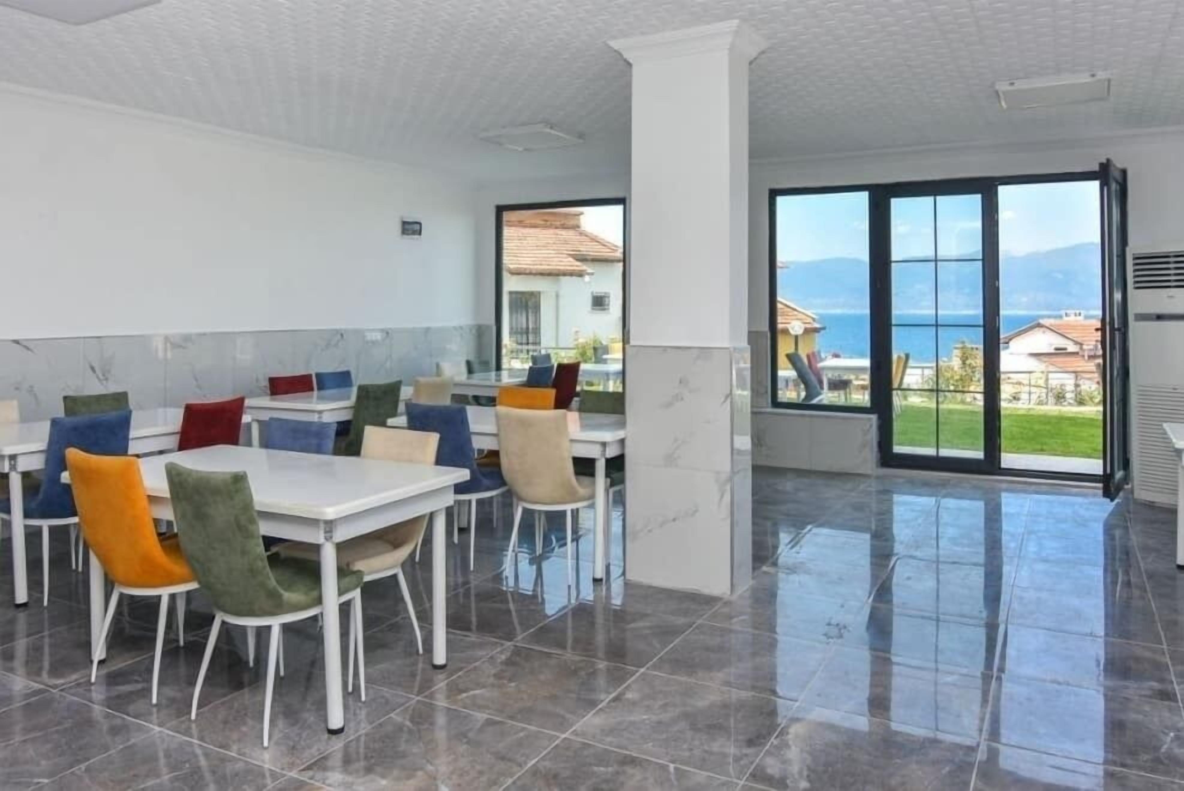 A spacious dining area with white tables and colorful chairs, large windows with a view of the sea and mountains, and a tiled floor.