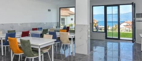 A spacious dining area with white tables and colorful chairs, large windows with a view of the sea and mountains, and a tiled floor.