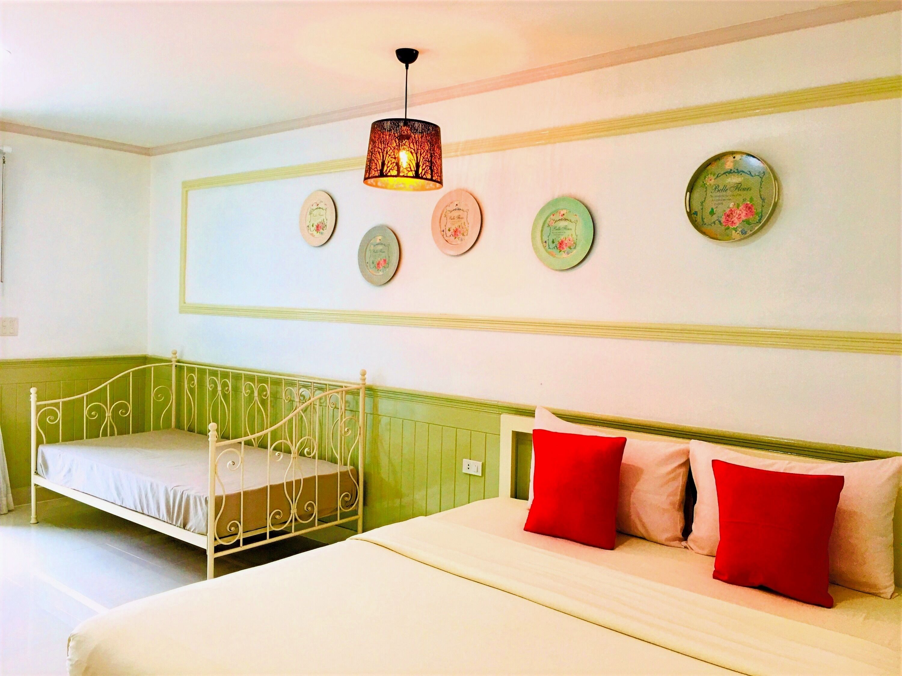 A room with a green accent wall, a white metal bed frame, a white mattress, and a red and white pillow. There is a wall-mounted light fixture above the bed.
