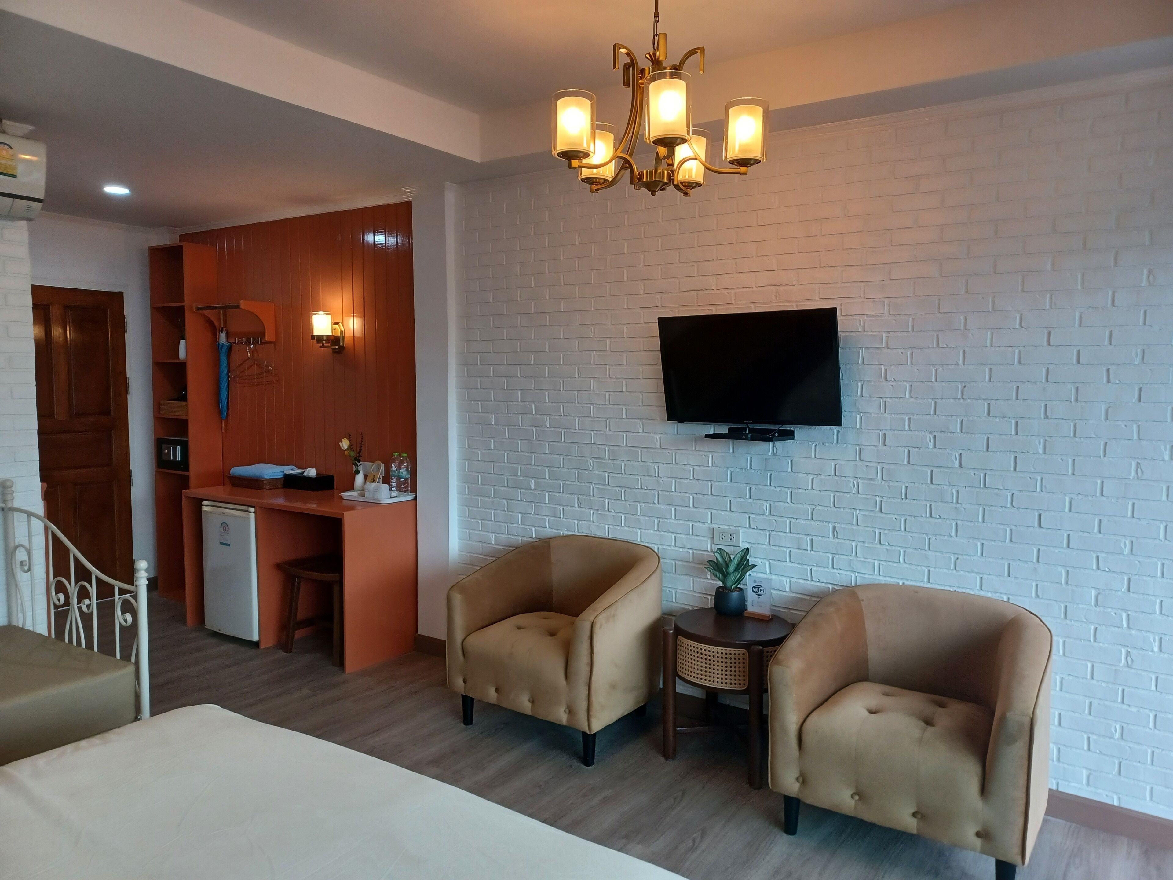 A hotel room with a bed, two armchairs, a small table, a TV mounted on a brick wall, a kitchenette, and a chandelier.