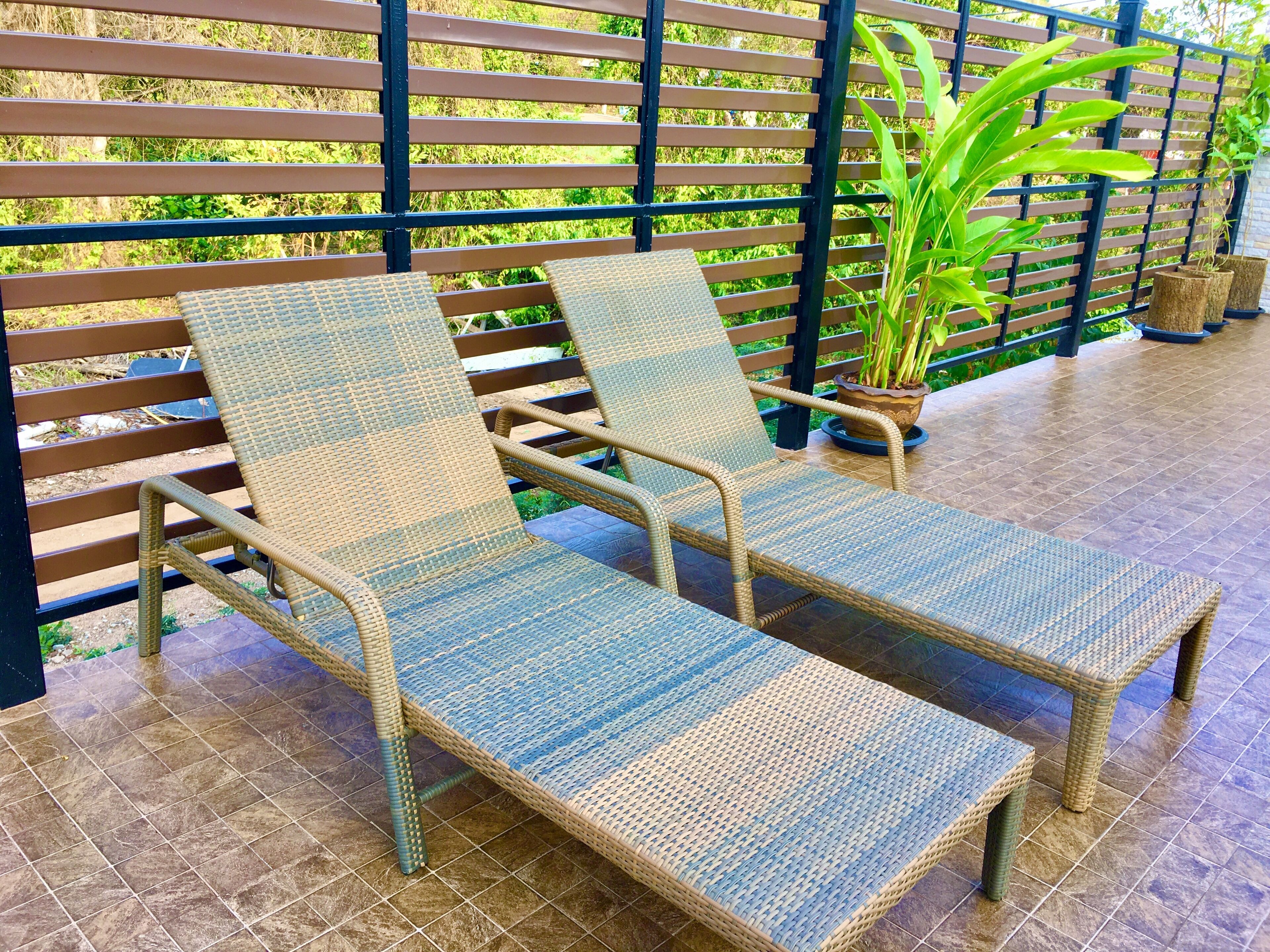 Ein Korbsessel mit einem blau-beige gestreiften Kissen, platziert auf einer gefliesten Terrasse mit Holzzaun und Blumentöpfen im Hintergrund.