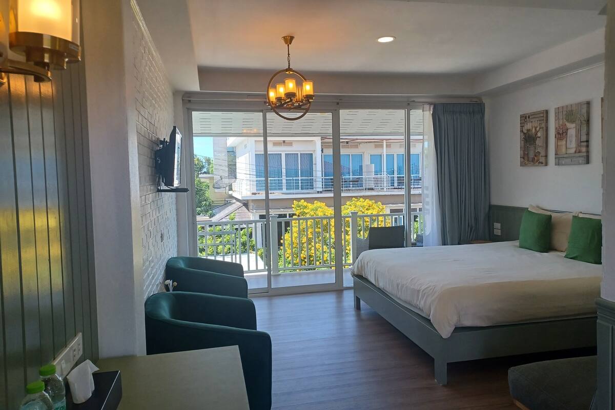 A hotel room with a bed, a desk, a chair, a balcony with a view of buildings and trees, and a chandelier.