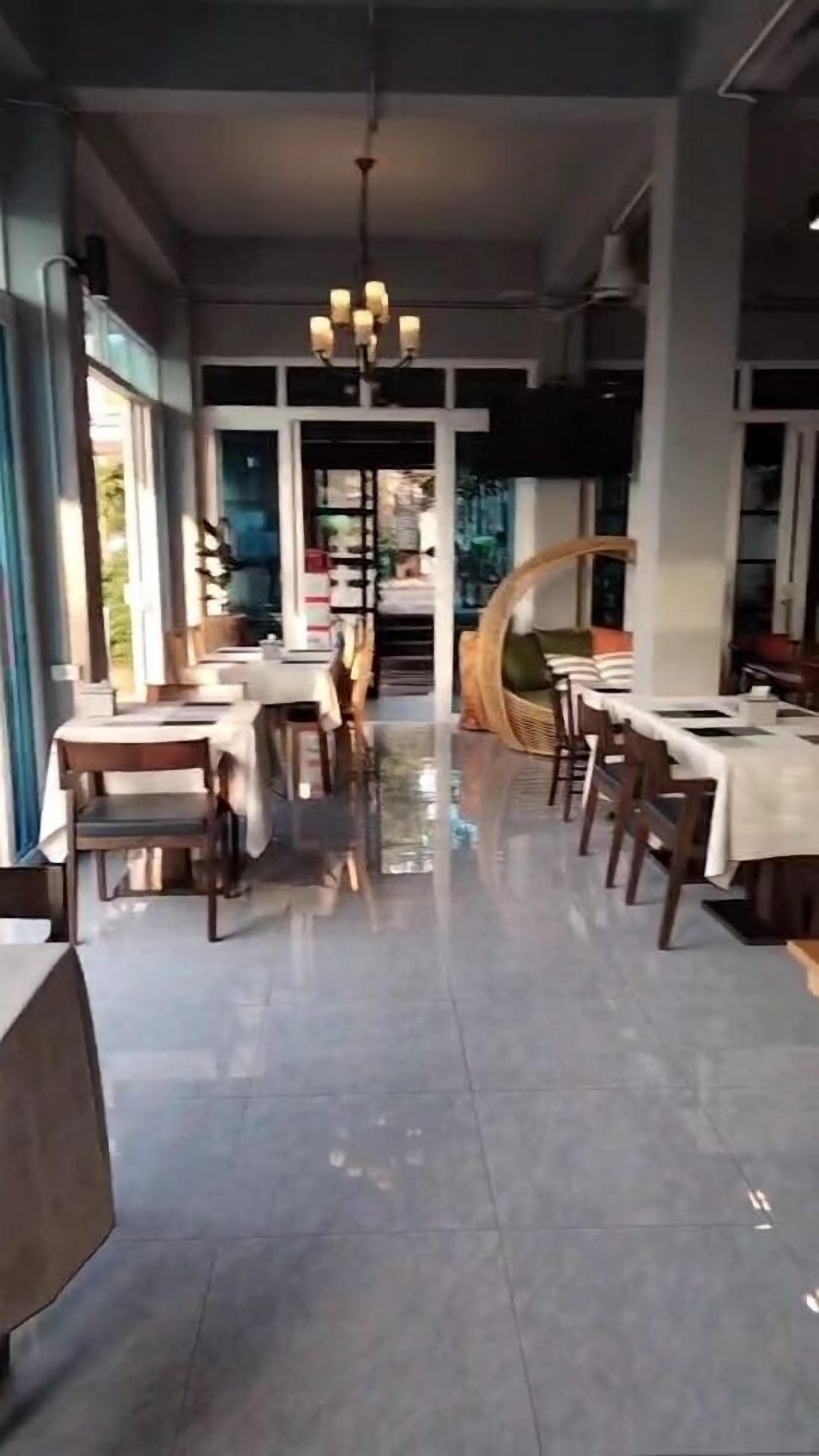 A restaurant with a modern interior, featuring a central chandelier, multiple tables, and a view through the glass doors.