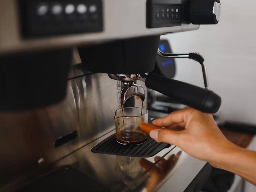 Una mano sosteniendo una taza debajo de una máquina de café, con una taza de vidrio transparente en el recipiente colector.