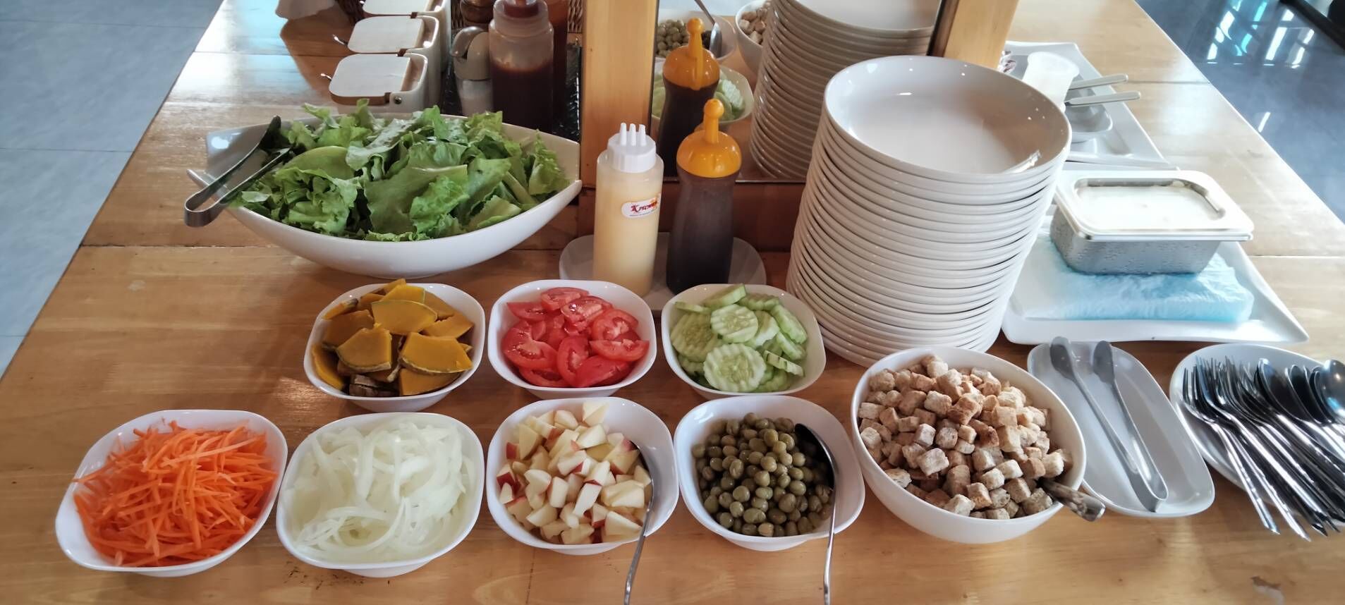 A table with various food items and condiments, including a bowl of salad, bowls of sliced vegetables, and a stack of plates.