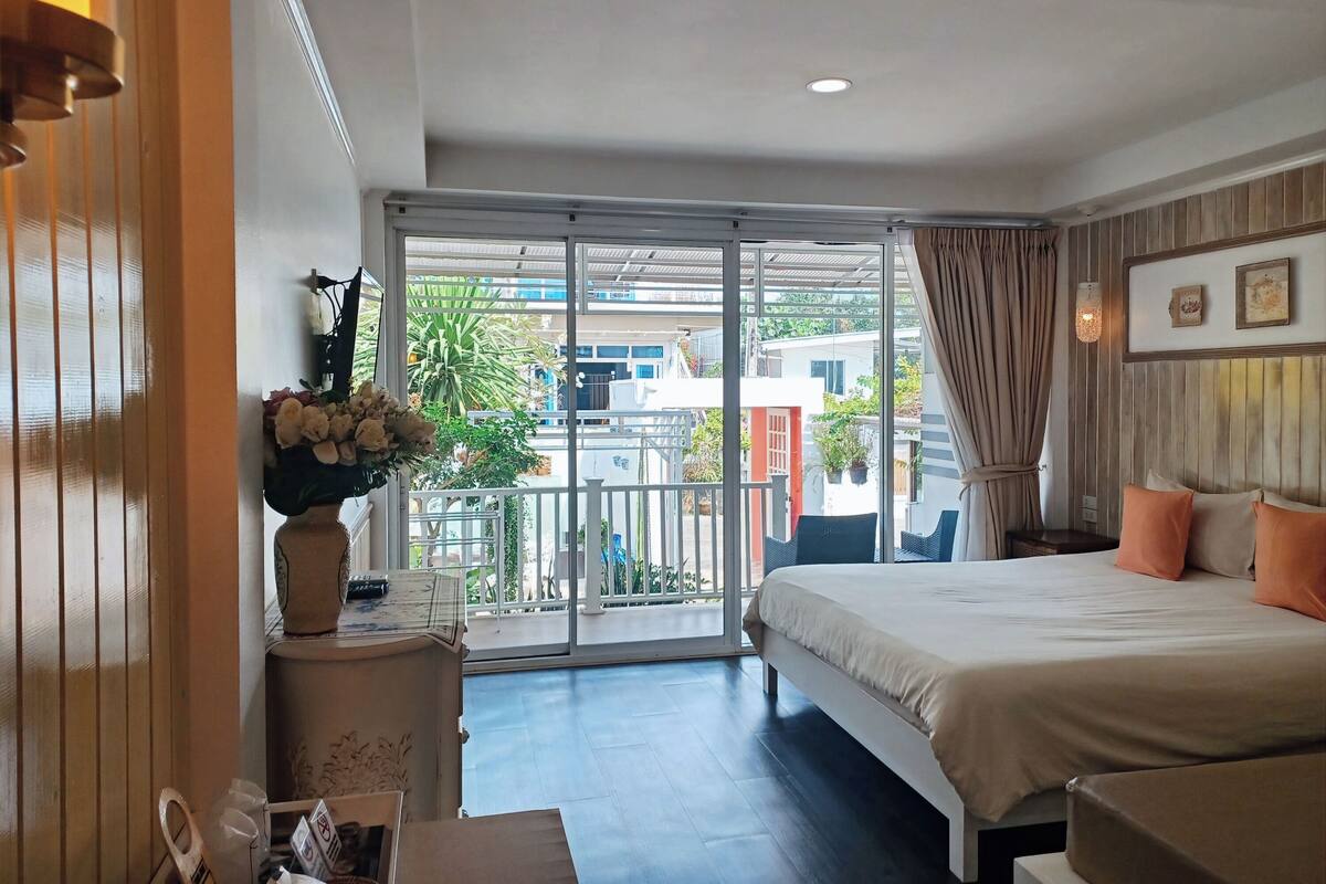 A hotel room with a bed, a nightstand, a vase with flowers, a window with a view of buildings and greenery, and a ceiling light.