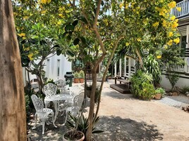 Un patio al aire libre con una mesa y sillas blancas, rodeado de macetas con plantas y árboles con flores amarillas.