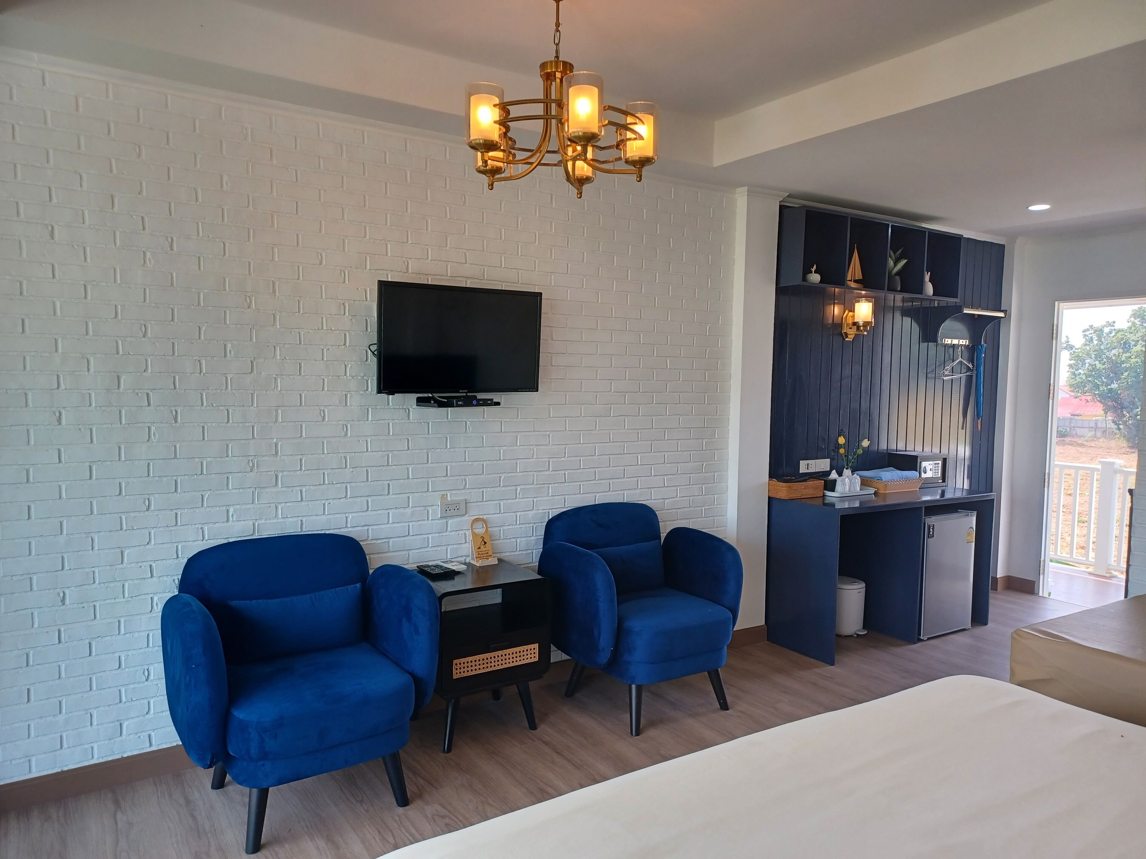 A modern room with a white brick wall, a flat-screen TV, two blue armchairs, a wooden table, and a chandelier.