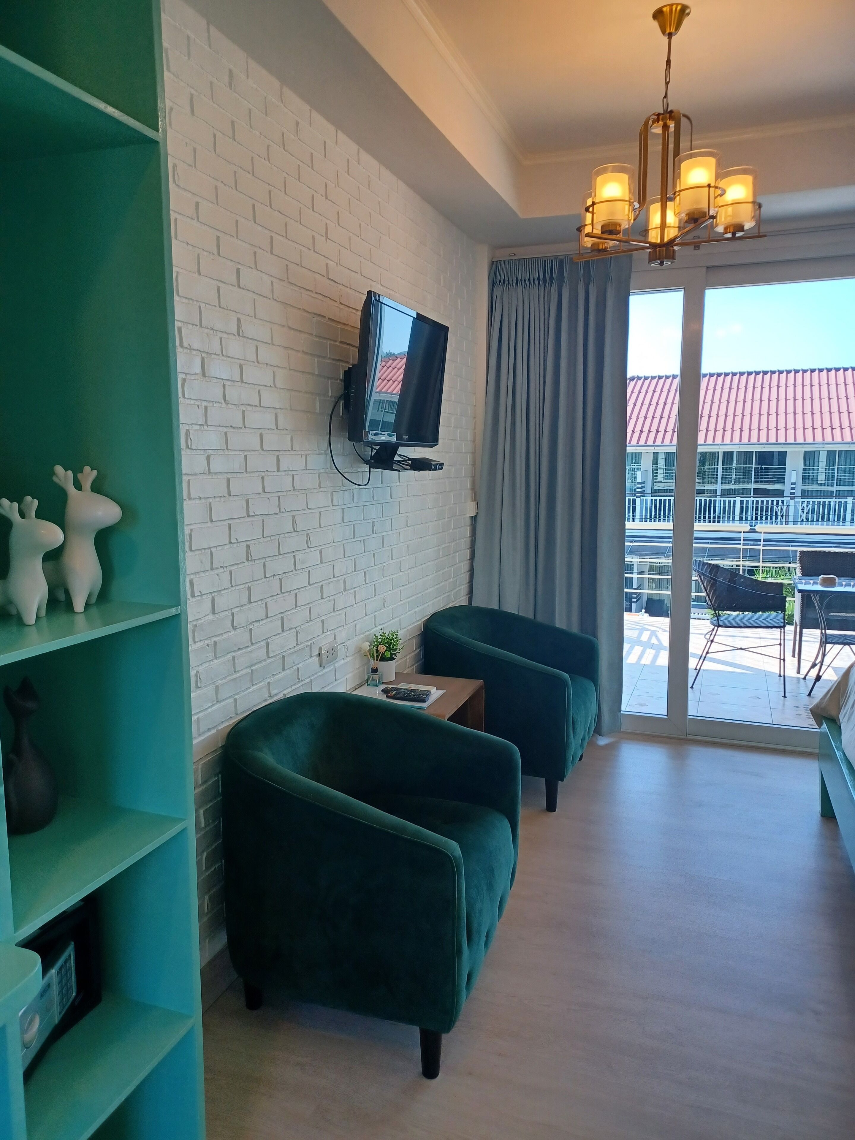 A modern living room with a white brick wall, a mounted TV, teal sofas, and a view of a building with a red roof through a glass door.