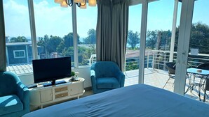 A hotel room with a large bed, a TV, a small table, a chair, and a balcony with a view of the city. - Koh Larn Riviera (Koh Lan)