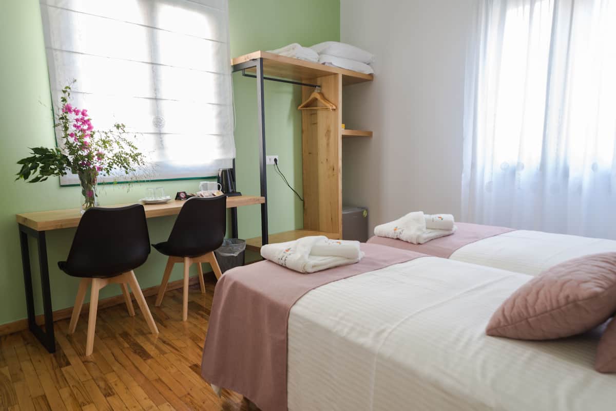 A bedroom with a bed, bedside table, two chairs, a desk, a hanging shelf, a window, and a potted plant.
