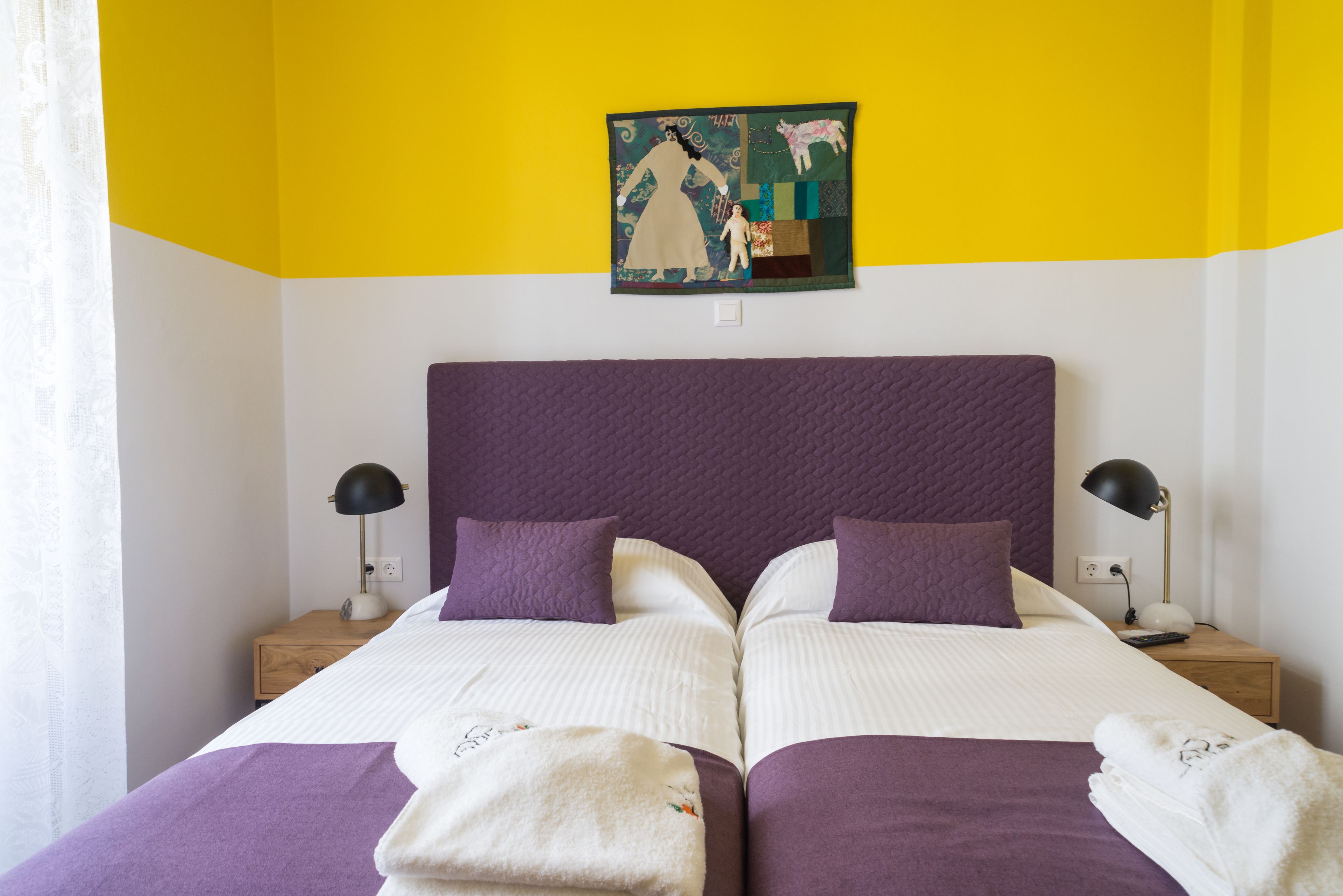 Una cama doble con cabecera morada, dos mesitas de noche con lámparas y un cuadro sobre la cama.