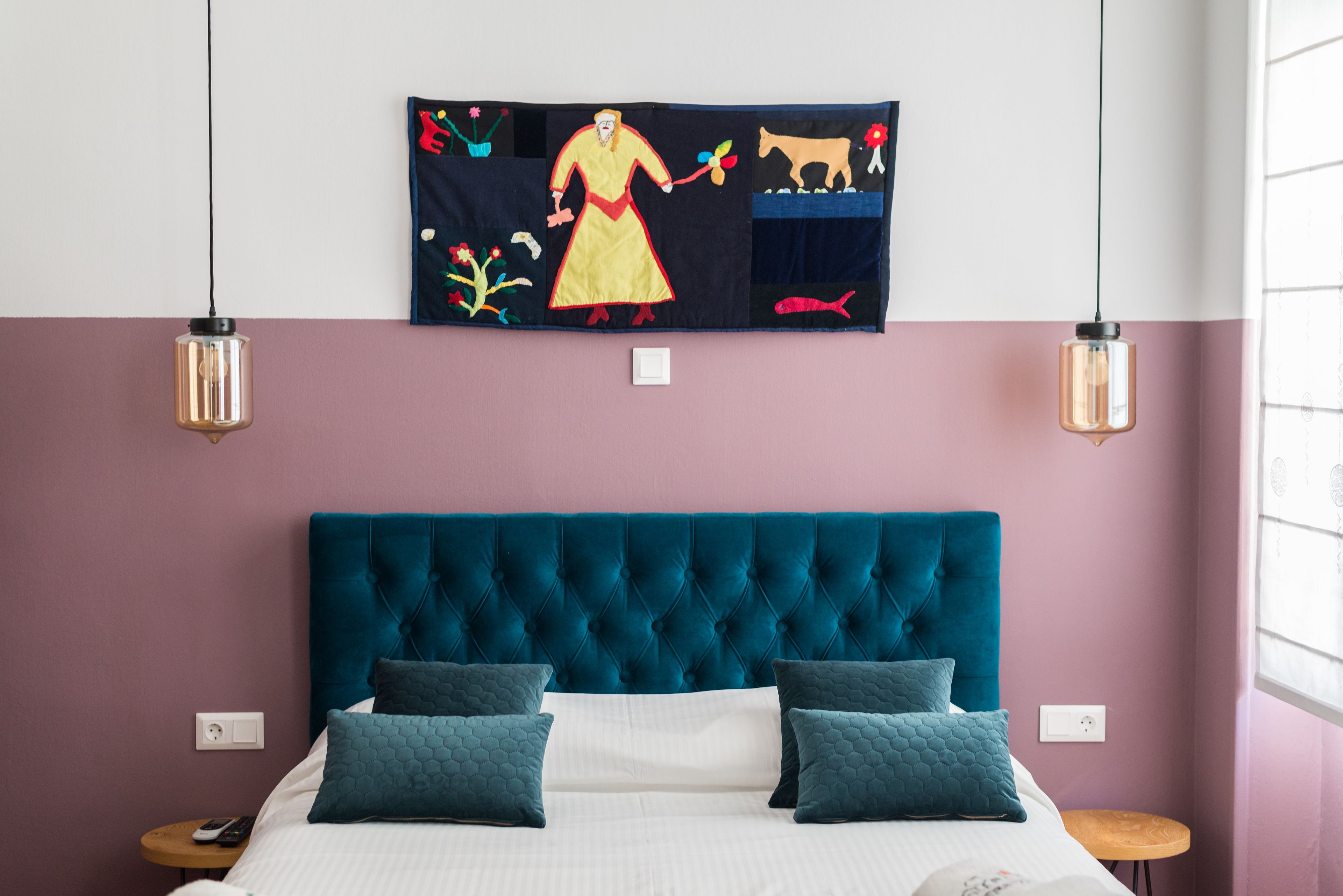 A bedroom with a teal upholstered headboard, a bed with white bedding and two teal pillows, a wall-mounted tapestry depicting a figure with animals, and two pendant lights.