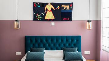 A bedroom with a teal upholstered headboard, a bed with white bedding and two teal pillows, a wall-mounted tapestry depicting a figure with animals, and two pendant lights.