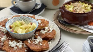 A breakfast setting with pancakes, apples, and feta cheese on a plate, a bowl of fruit, a cup of coffee, and a vase with yellow flowers. - Neratzia Rooms (Athens)