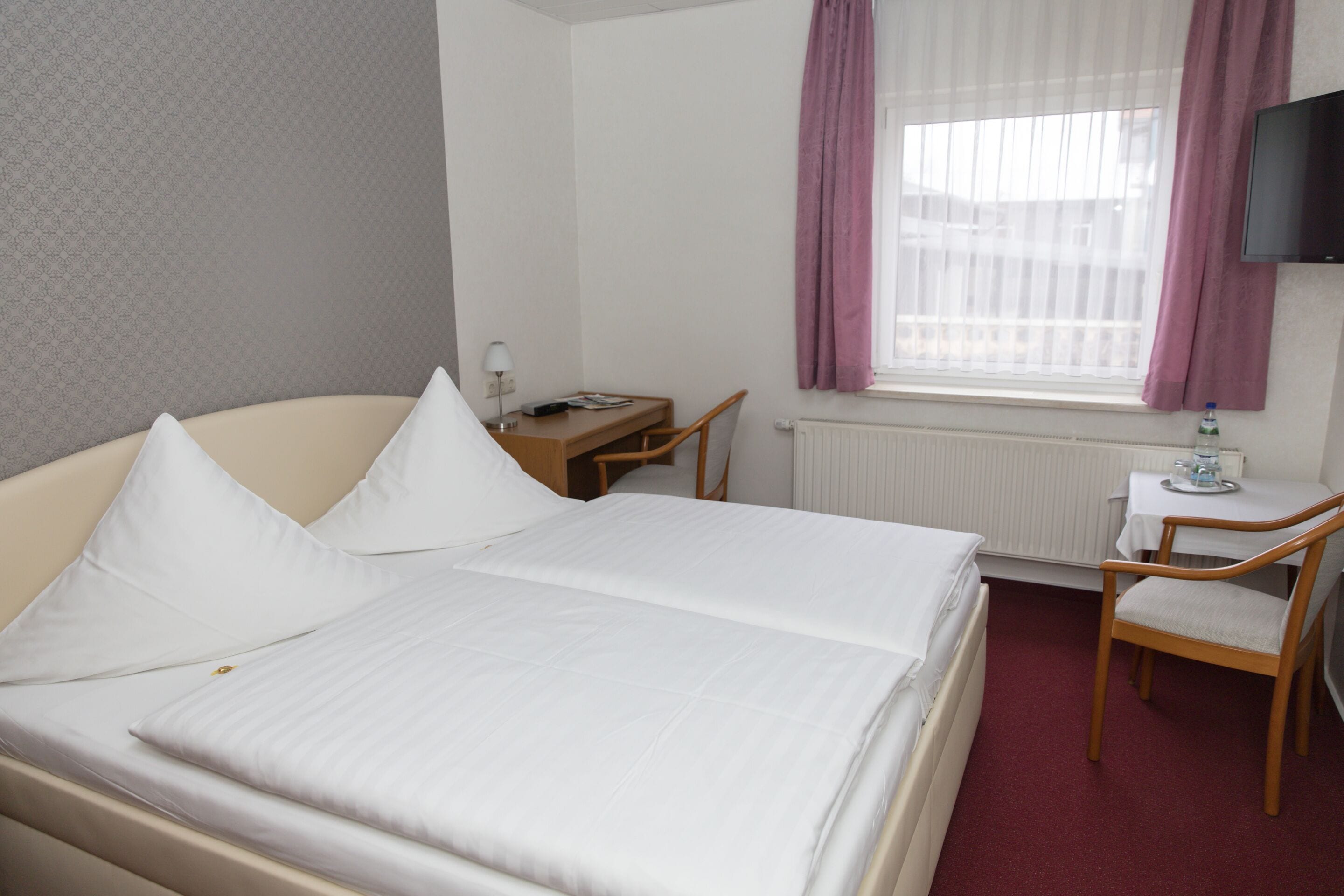 A hotel room with a bed, a desk, a chair, a TV, and a window with curtains.