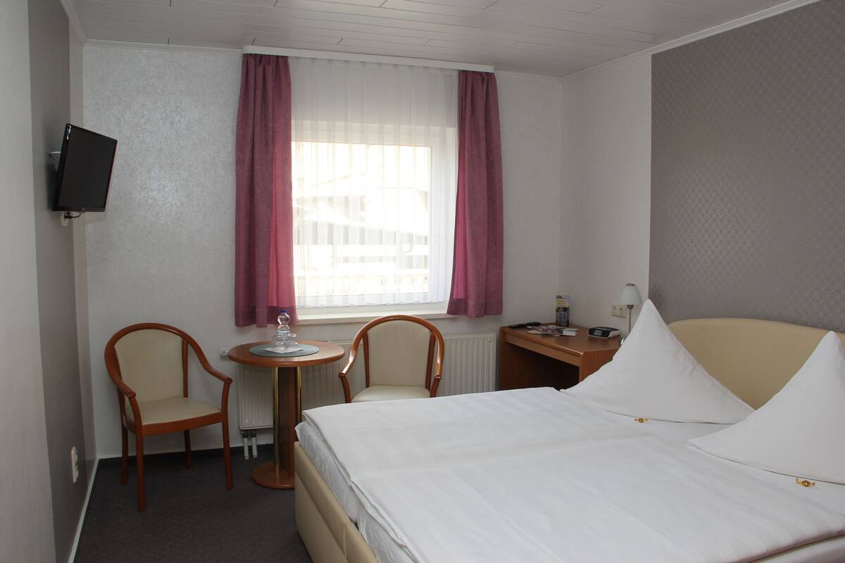 A hotel room with a bed, two chairs, a small table, a TV, and a window with curtains.