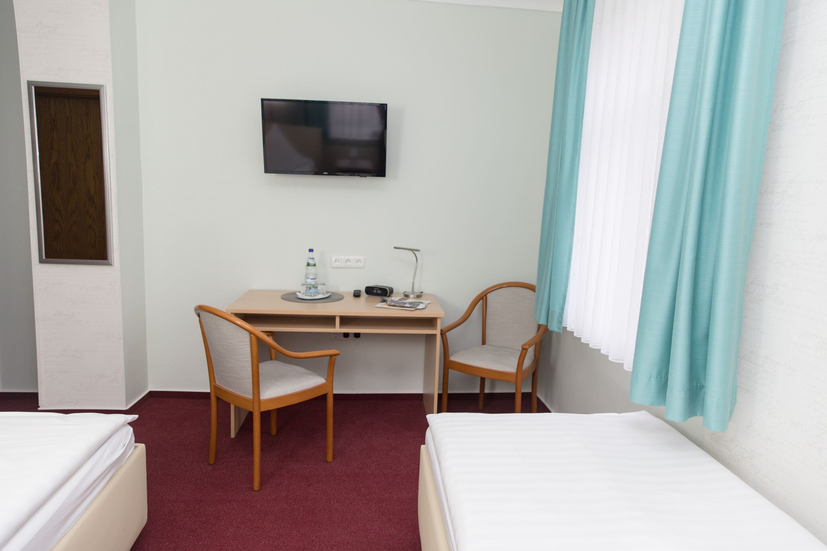 A hotel room with two single beds, a desk with a chair, a television, and a window with curtains.