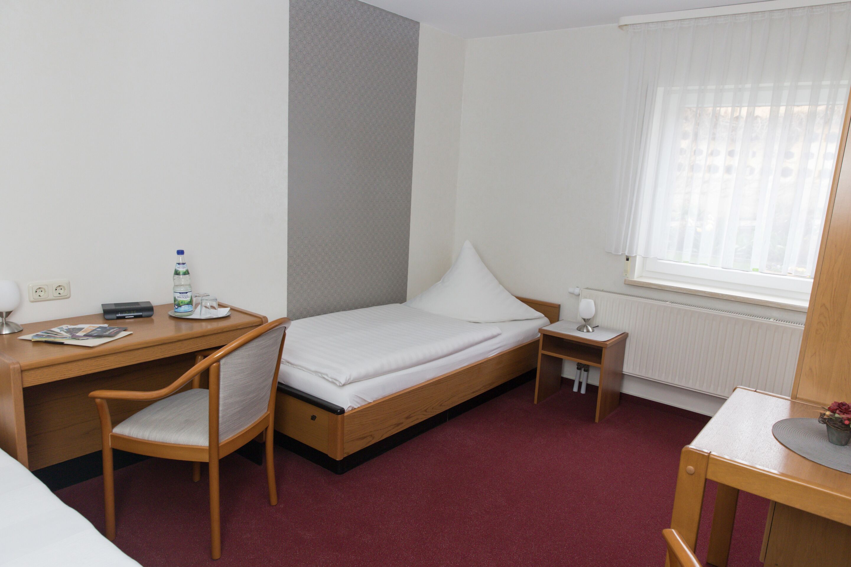 A small, neatly arranged hotel room with a single bed, a desk, a chair, a small table, and a window with curtains.