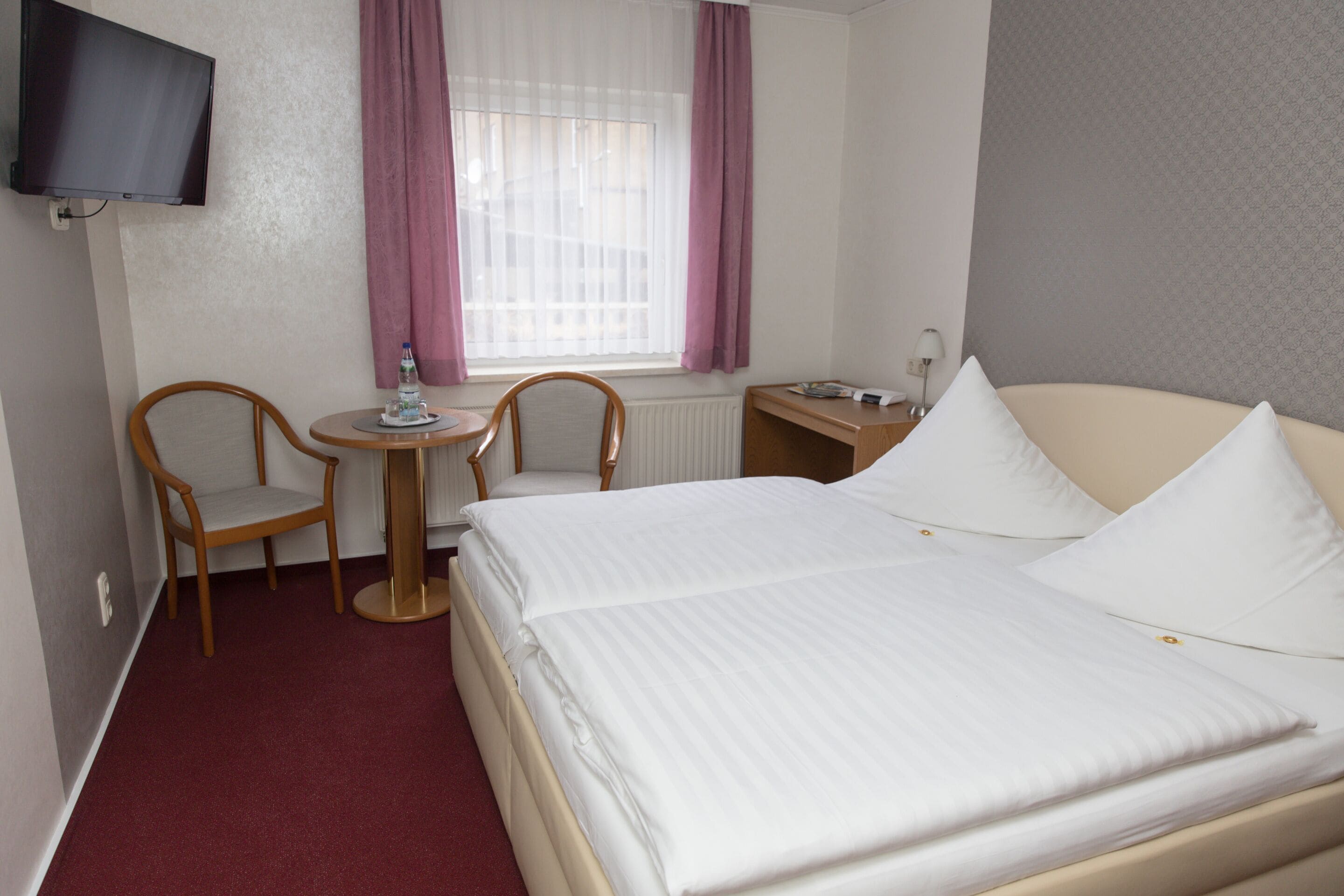 Ein Hotelzimmer mit einem Bett, zwei Sesseln, einem kleinen Tisch, einem an der Wand befestigten Fernseher und einem fenster mit Vorhängen.