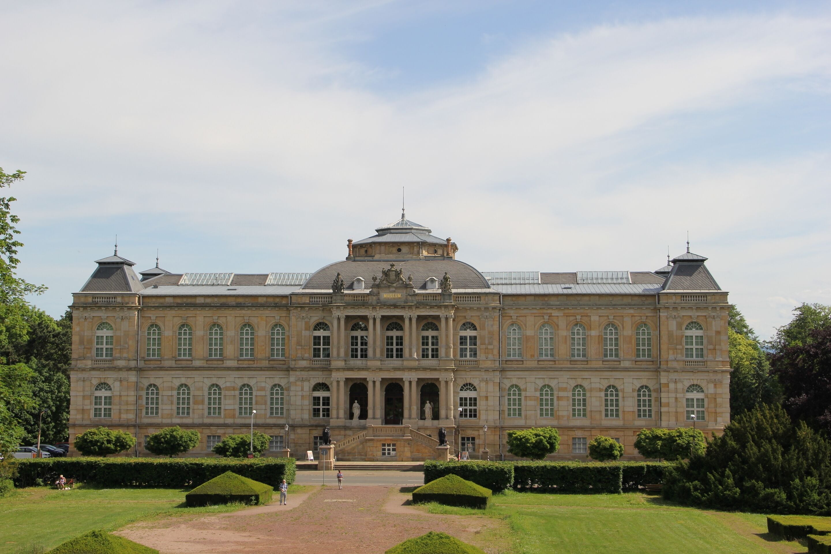 A large, historic building with a central dome and symmetrical wings, surrounded by well-manicured gardens.
