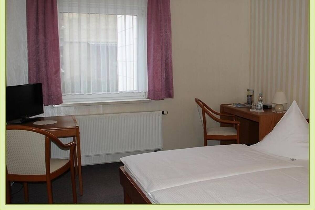 A hotel room with a bed, a desk with a chair, a television, and a window with curtains.