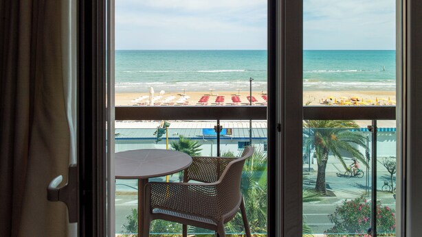 Un balcón con sillas de mimbre y una mesa, con vistas a una playa con sombrillas y un cielo despejado.
