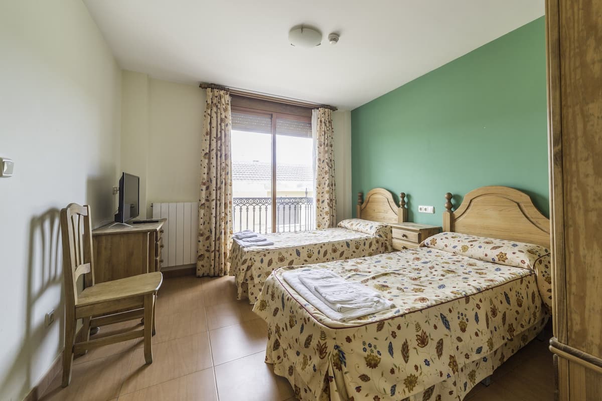 Una habitación con dos camas, un escritorio con computadora, una silla y un ventanal con cortinas.