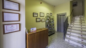 Uma área de recepção com balcão de madeira, certificados emoldurados na parede, uma vitrine de vidro e escada com corrimão de metal.