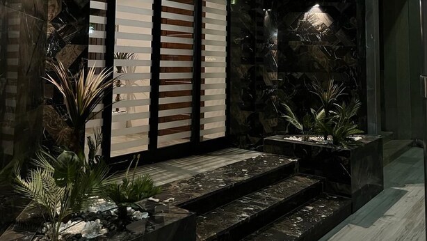 Fachada de edificio moderno de piedra, con una escalera que conduce a una puerta y un pequeño jardín con plantas.