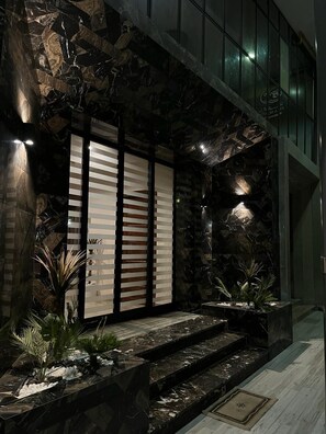 A modern building exterior with a stone facade, a set of stairs leading to a door, and a small garden area with plants. - Pacha Hotel (Sfax)