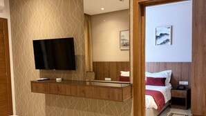 Una habitación de hotel con una cama, un televisor montado en un estante de madera, una mesita de noche con una lámpara y una puerta que da a otra habitación.