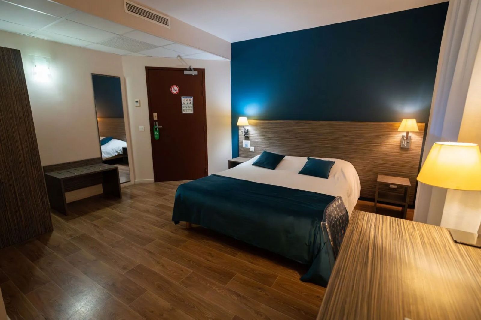 Habitación de hotel con cama, lámparas de noche, un banco y una puerta con un cartel de prohibido fumar.
