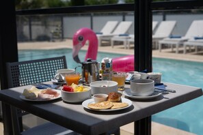 Una mesa de desayuno con diversos alimentos y bebidas, con vista a una piscina que tiene sillas de descanso y un flotador con forma de flamenco rosa.